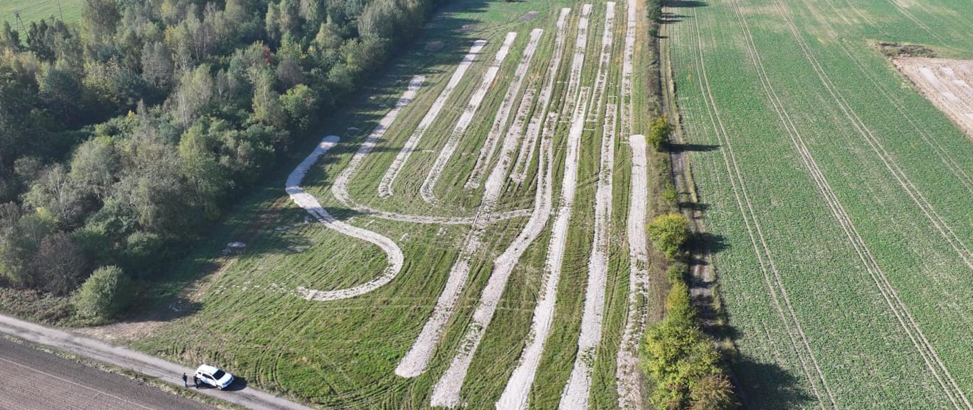 Zdjęcie pola z wysokości kilkudziesięciu metrów w pobliżu lasu, na którym widoczne są równoległe pasy białych osadów ściekowych rozmieszczone na zielonej powierzchni trawy. W dolnym rogu widać drogę z zaparkowanym samochodem.