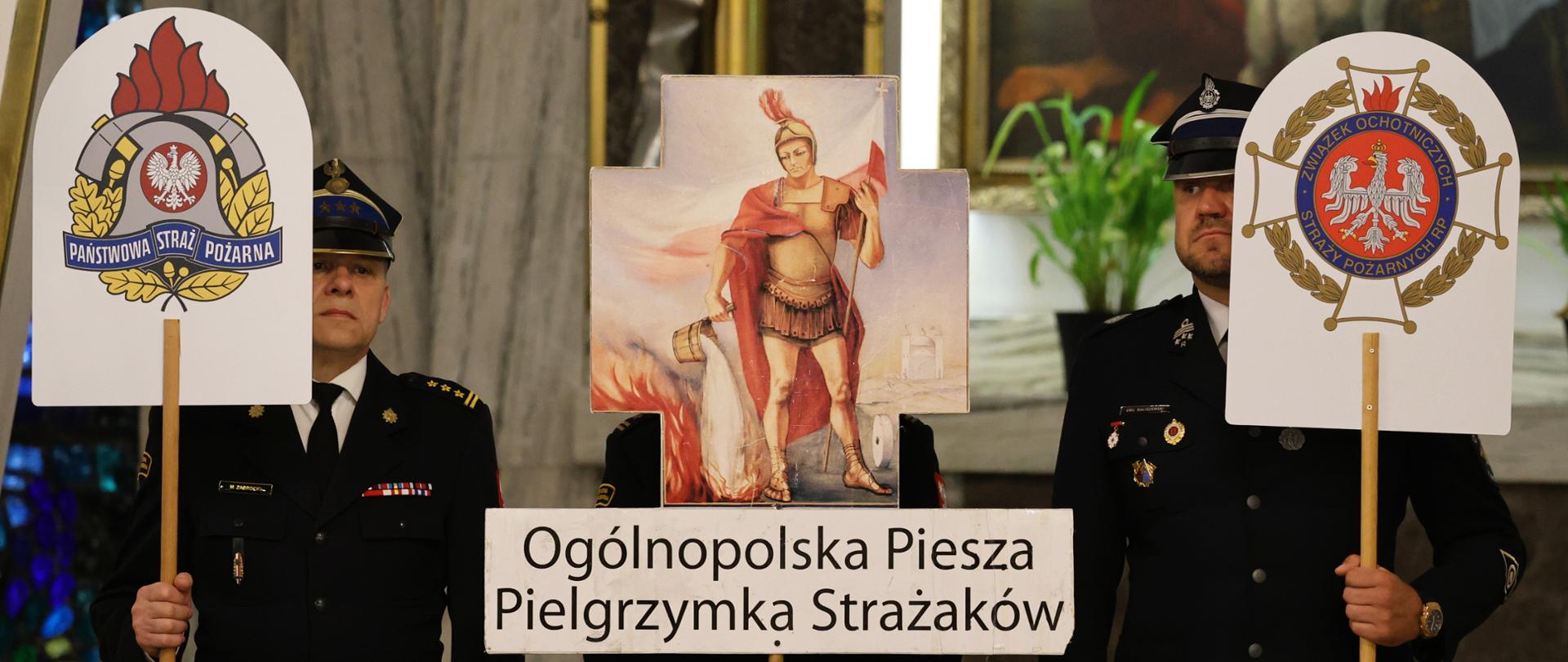 Strażacy Państwowej Straży Pożarnej i Ochotniczych Straży Pożarnych w umundurowaniu galowym stoją w Sanktuarium Matki Boskiej Ostrobramskiej w Warszawie przy znakach pielgrzymkowych XX Ogólnopolskiej Pieszej Pielgrzymki Strażaków na Jasną Górę. W centralnej części widoczny jest symbol św. Floriana oraz tablica z nazwą pielgrzymki. 
