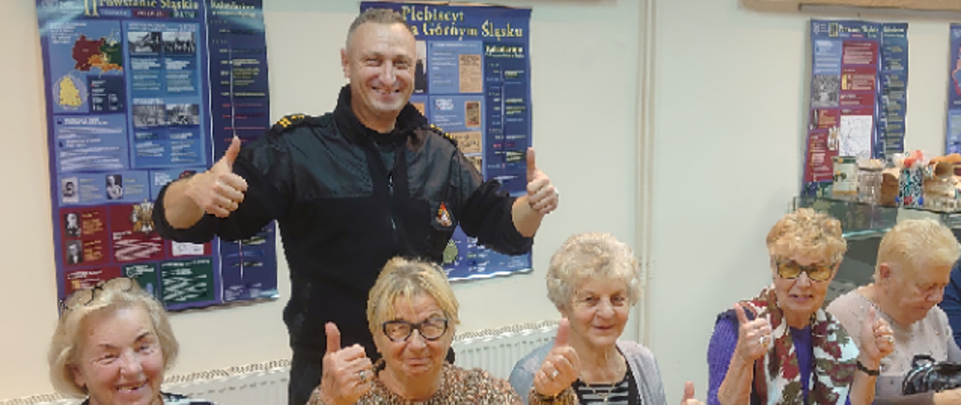 Na zdjęciu znajdują się kobiety siedzące przy stolikach w pomieszczeniu, na stolikach są ustawione butelki z wodą w tle stoi strażak ubrany w ubiór koszarowy