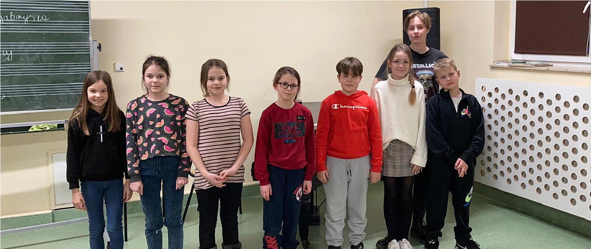 Grupa uczniów, pięć dziewczynek i trzech chłopców pozuje do zdjęcia, dzieci stoją w pomieszczeniu, na podłodze koloru seledynowego, za nimi z lewej strony przy ścianie koloru słomkowego stoi tablica szkolna do pisania nut, pośrodku za nimi widać fortepian, z prawej strony widać okno zasłonięte roletą a pod nim kaloryfer osłonięty białą płytą z wyciętymi otworami.