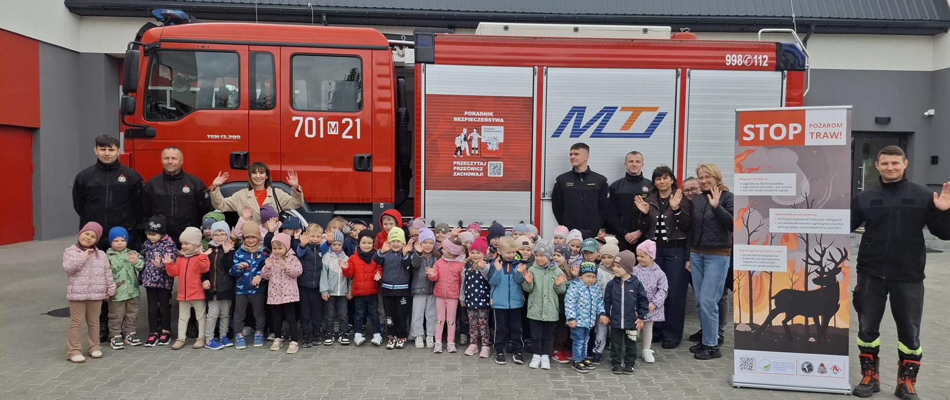 Grupa kilkudziesięciu dzieci z opiekunami oraz strażakami pozuje do wspólnego zdjęcia przed czerwonym wozem strażackim zaparkowanym przed nowoczesnym budynkiem straży pożarnej. Dzieci ubrane są w kolorowe kurtki i czapki, wiele z nich macha do aparatu. Po prawej stronie stoi baner edukacyjny dotyczący zapobiegania pożarom traw z dużym napisem „STOP pożarom traw!”. Strażacy ubrani są w ciemne mundury, a w tle widoczna jest szara elewacja budynku i pochmurne niebo.