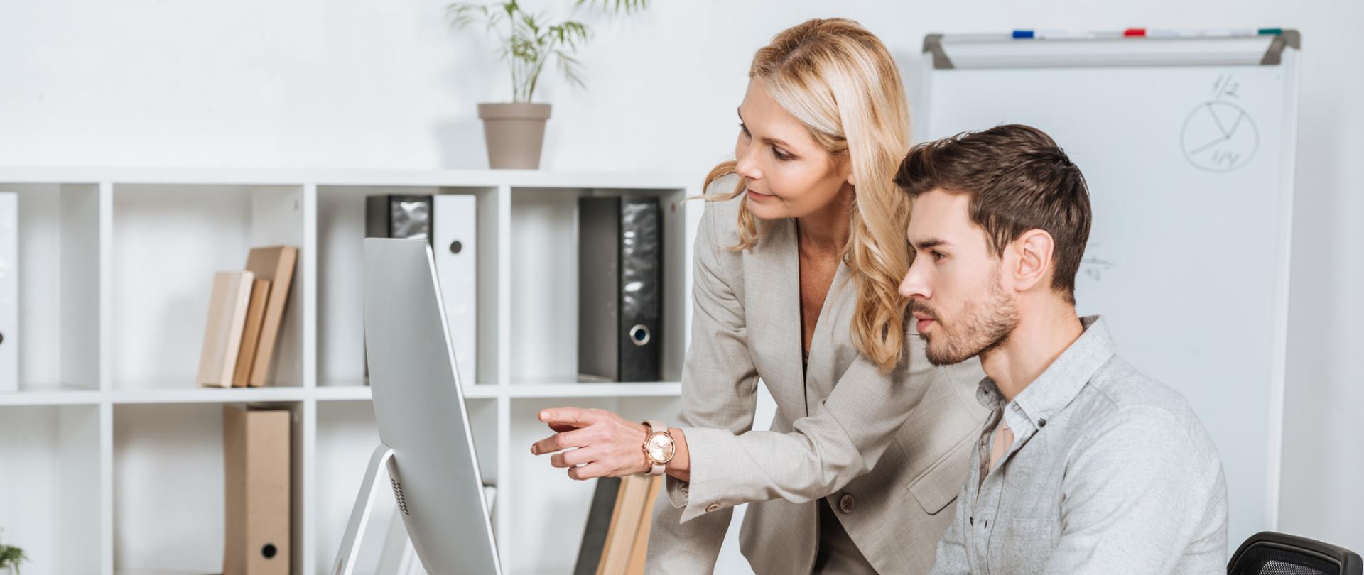 Na zdjęciu jest kobieta i mężczyzna będący przed komputerem. W tle widać biuro.