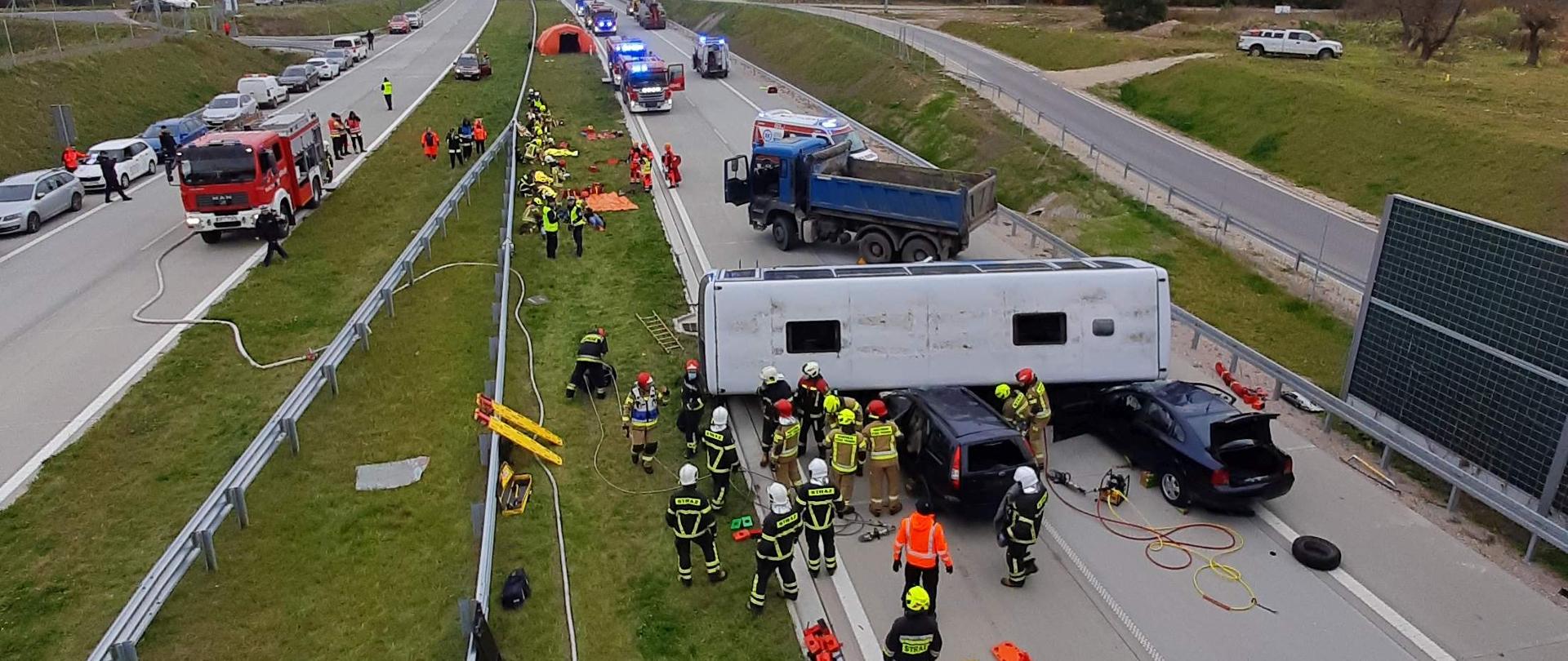 Autobus leży na boku podparty na 2 samochodach osobowych. Wokół widocznych kilkunastu strażaków ze sprzętem. W głębi rozstawiony namiot dla poszkodowanych.