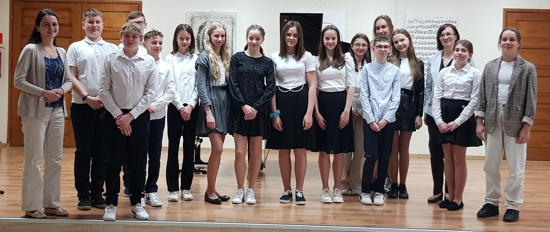 Fotografia przedstawia uczestników audycji fortepianu dodatkowego i ich nauczycieli. Wszyscy stoją na scenie auli PSM I st. w Grajewie. W tle fortepian, grafiki przedstawiające partytury i postać Jana Sebastian Bacha, oraz drzwi w kolorze brązowym. 