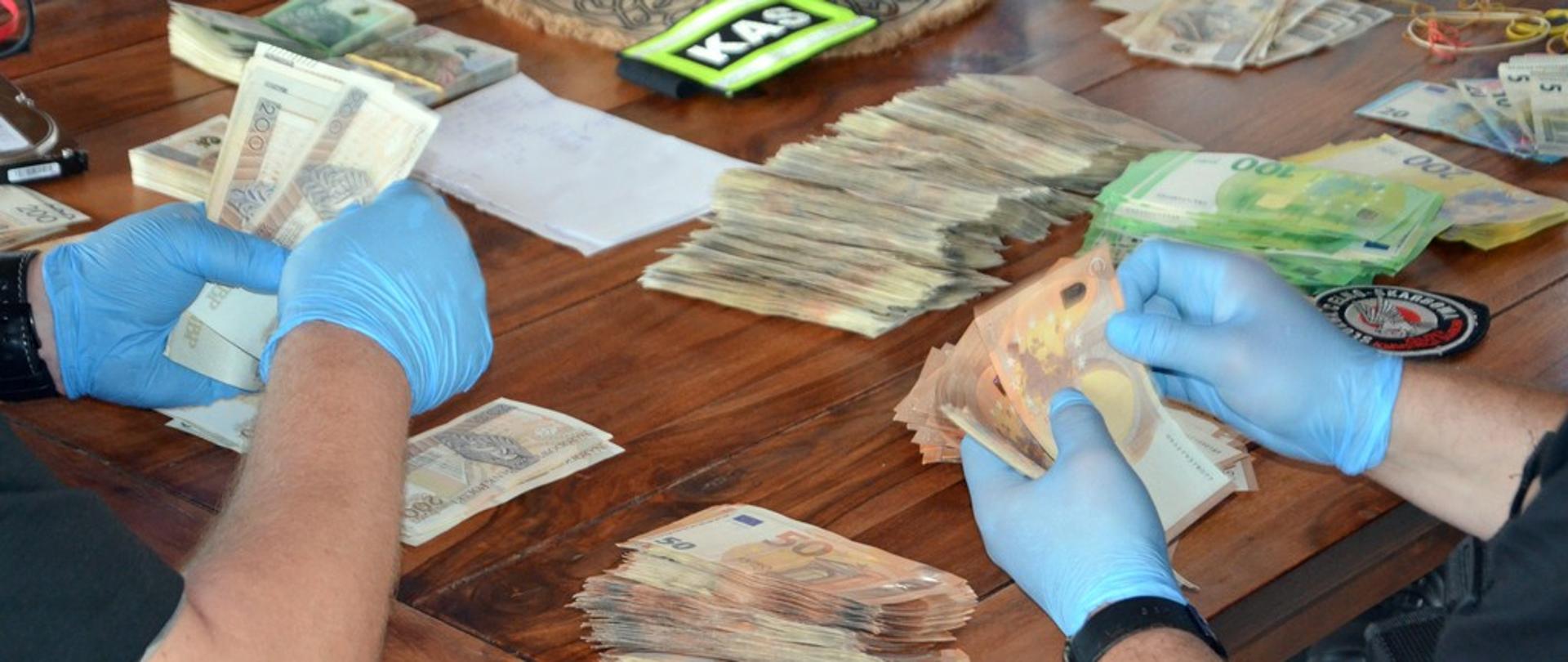 Osoby w rękawiczkach liczą i segregują banknoty różnych walut na drewnianym stole.