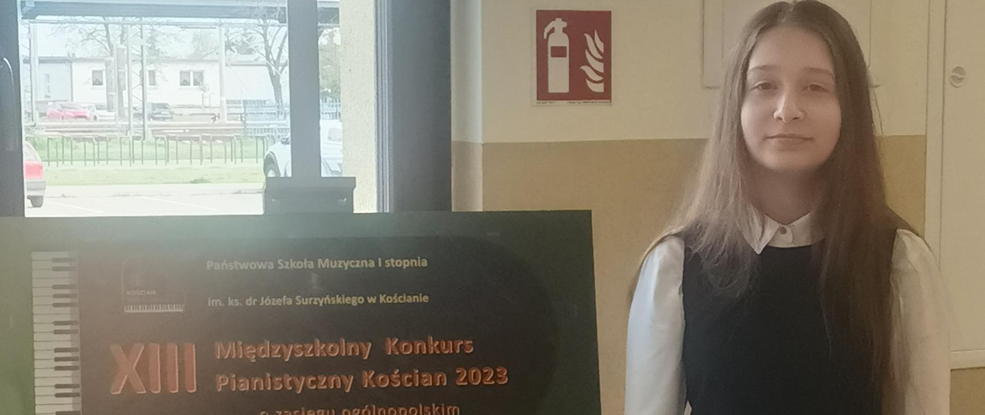 Zdjęcie przedstawiające uczennice Karolinę Podborę stojącą obok plakatu konkursowego stojącego na sztaludze. Zdjęcie wykonane w budynku szkoły. 