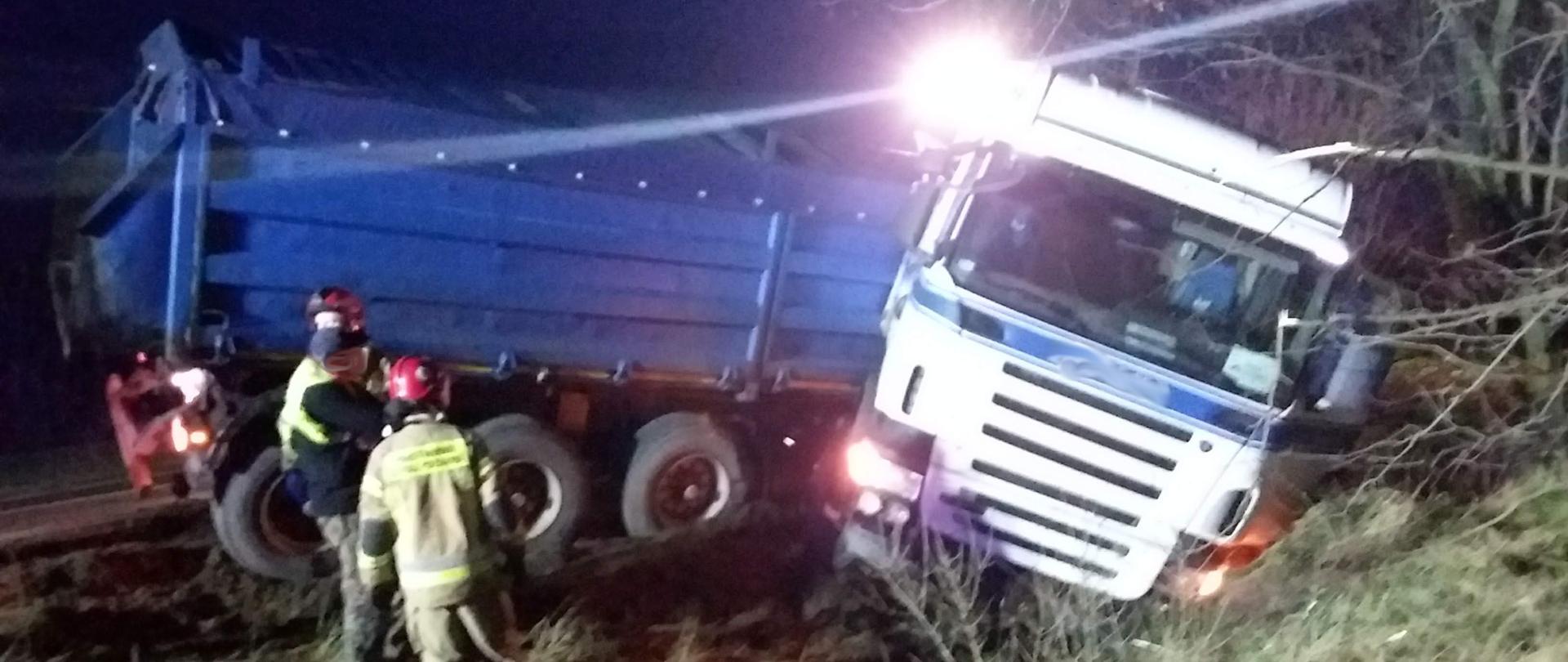 Ciężarówka z niebieską naczepą leży w rowie. Część naczepy na drodze. Wokół strażacy uczestniczący w działaniach ratowniczych.