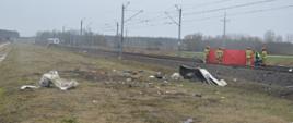 Wypadek śmiertelny na przejeździe kolejowym. Okolice miejscowości Kąty (gm. Rawicz). Na torowisku rozstawiony jest czerwony parawan ochronny, przy pomocy którego zabezpieczone zostało ciało ofiary wypadku. Przed nasypem, w rowie - roztrzaskana kabina ciężarówki. W oddali pociąg uczestniczący w zdarzeniu.