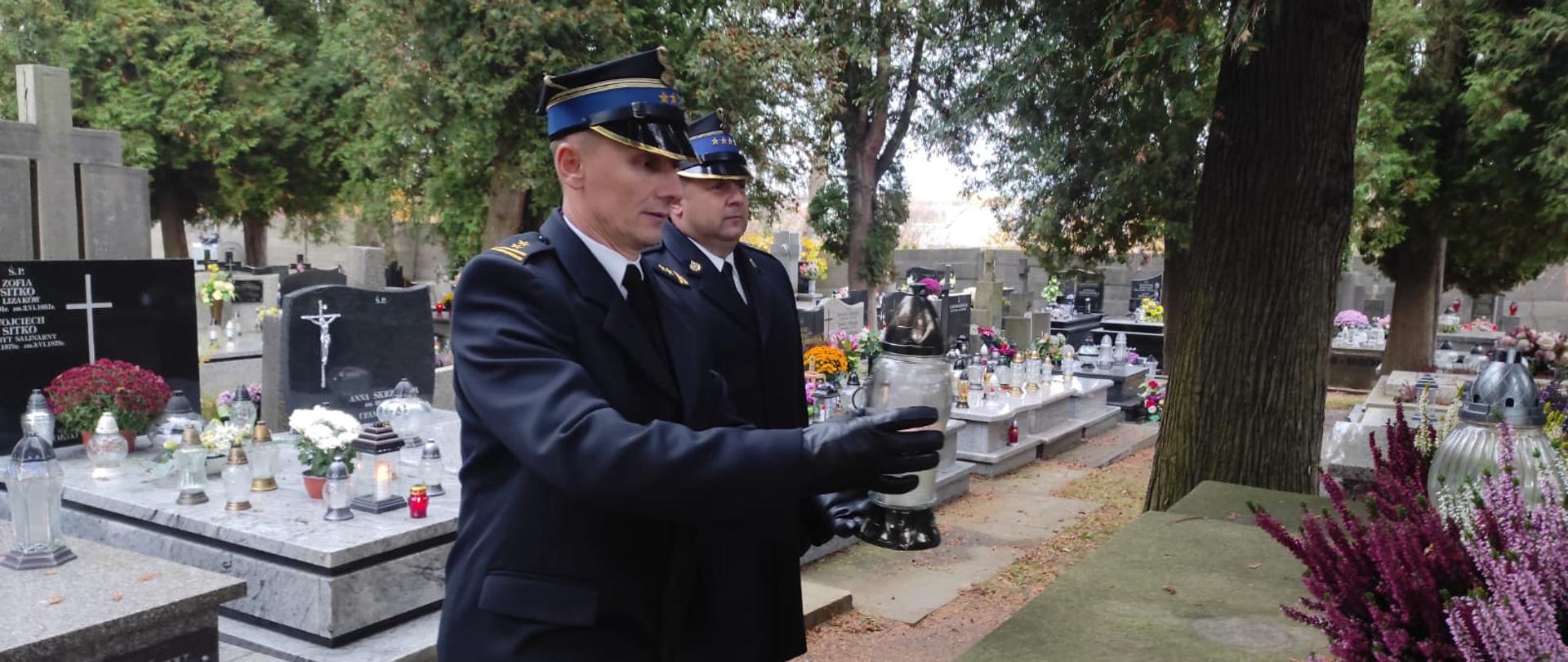 Dwóch strażaków w eleganckich płaszczach znajduje się na cmentarzu pośród nagrobków. Jeden strażak trzyma znicz w ręce. Drugi ma postawę zasadniczą.