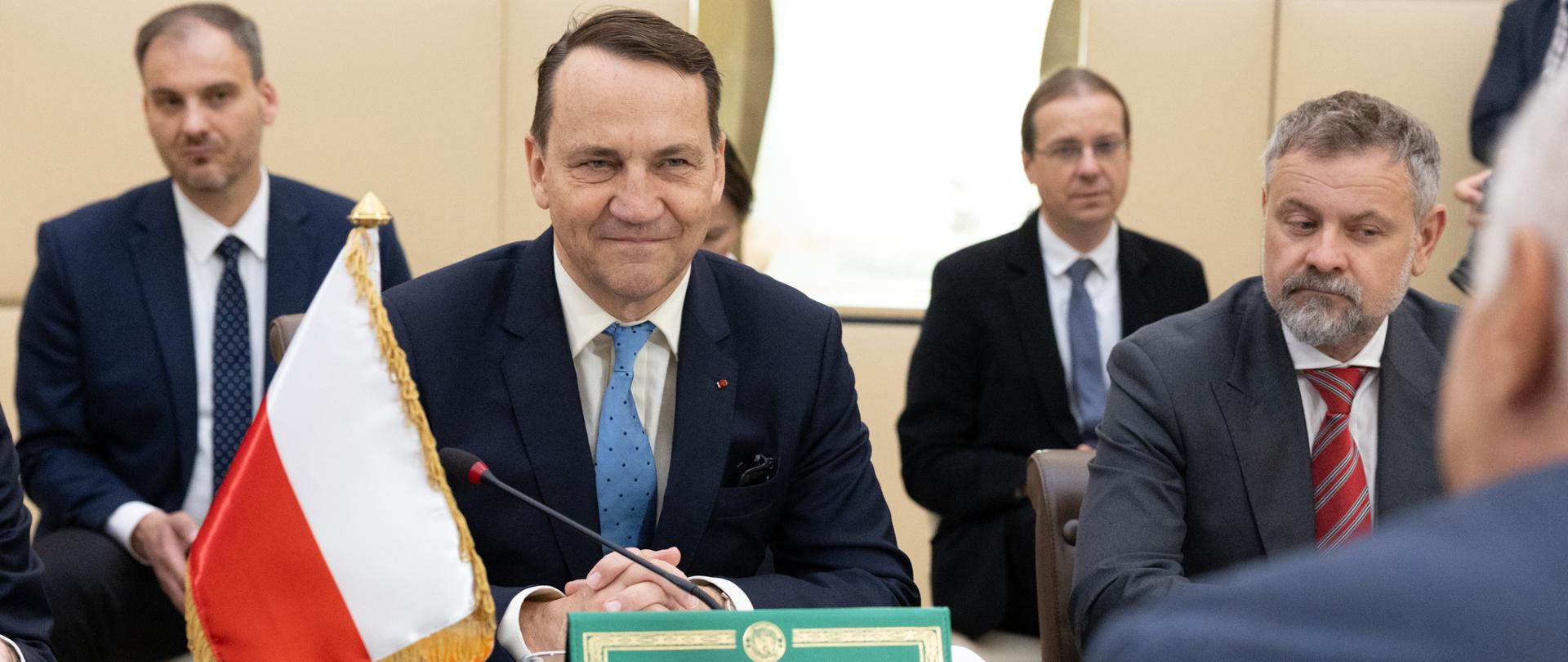 In the foreground, Deputy Prime Minister, Minister of Foreign Affairs Radosław Sikorski in a navy suit with a light blue tie sits with his hands clasped. On the table there is a Polish flag, a microphone, glasses of water, and documents, while a person in the foreground gestures with their hand, seen from behind.