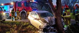 Na zdjęciu widać rozbity samochód osobowy, po uderzeniu w drzewo. Przy wraku widać strażaków. Na drugim planie dwa samochody straży pożarnej z włączonymi sygnałami błyskowymi.