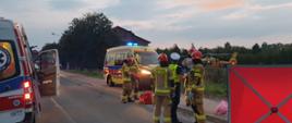 Widać drogę wojewódzką. Na środku służby ratunkowe. Pojazdy służb ratunkowych. Zdjęcie wykonane wieczorem. W tle zabudowania. Po prawej stronie śmigłowiec LPR. Po lewej ZRM. Na pierwszym planie asfalt.