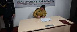 Strażak w piaskowym mundurze podpisuje akt ślubowania przy biurku na tle tablicy z logo Komendy Powiatowej PSP w Wodzisławiu Śląskim.
