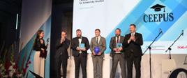 Pod wielkim ściennym ekranem z napisem CEEPUS Week stoi pięciu mężczyzn w garniturach, trzymają małe czworokątne niebieskie przedmioty, z boku przy mikrofonie stoi kobieta.