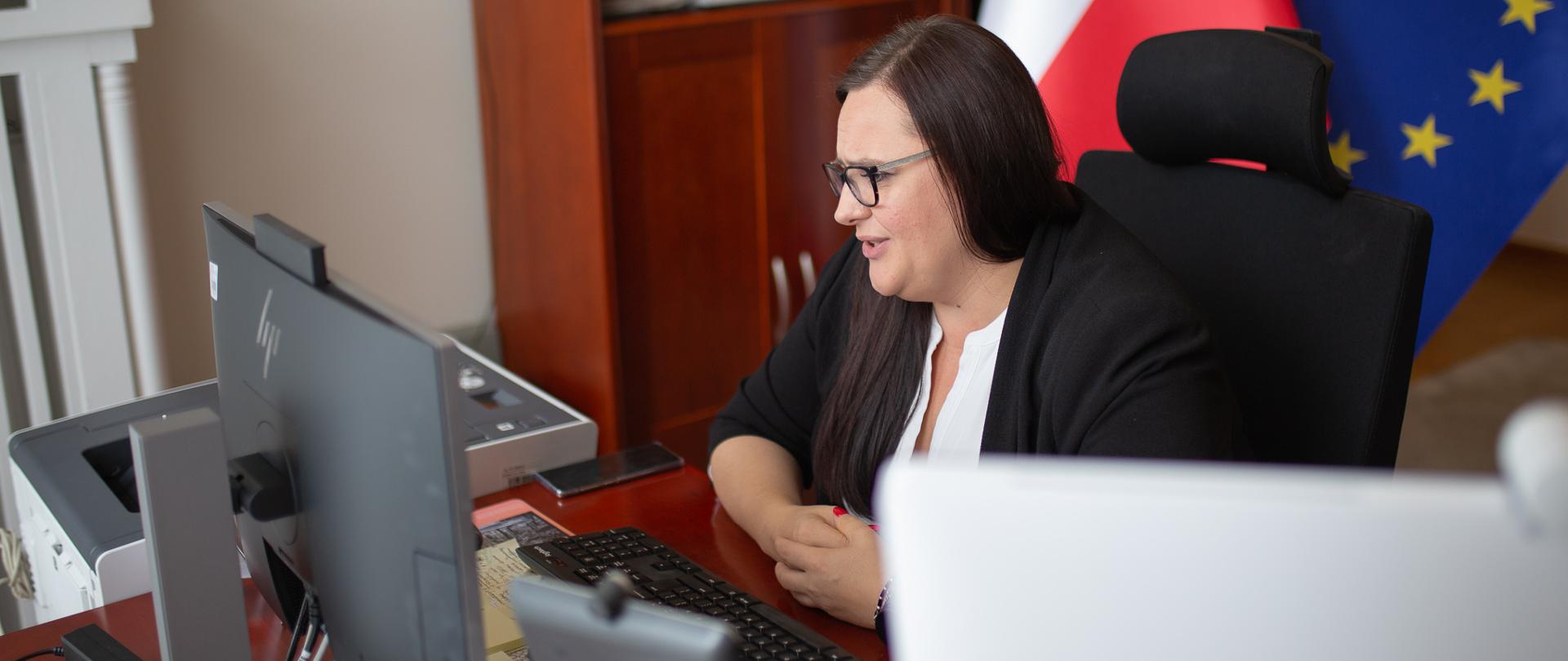Minister Małgorzata Jarosińska-Jedynak w służbowym pokoju przy biurku prowadzi zdalną rozmowę przez komputer
