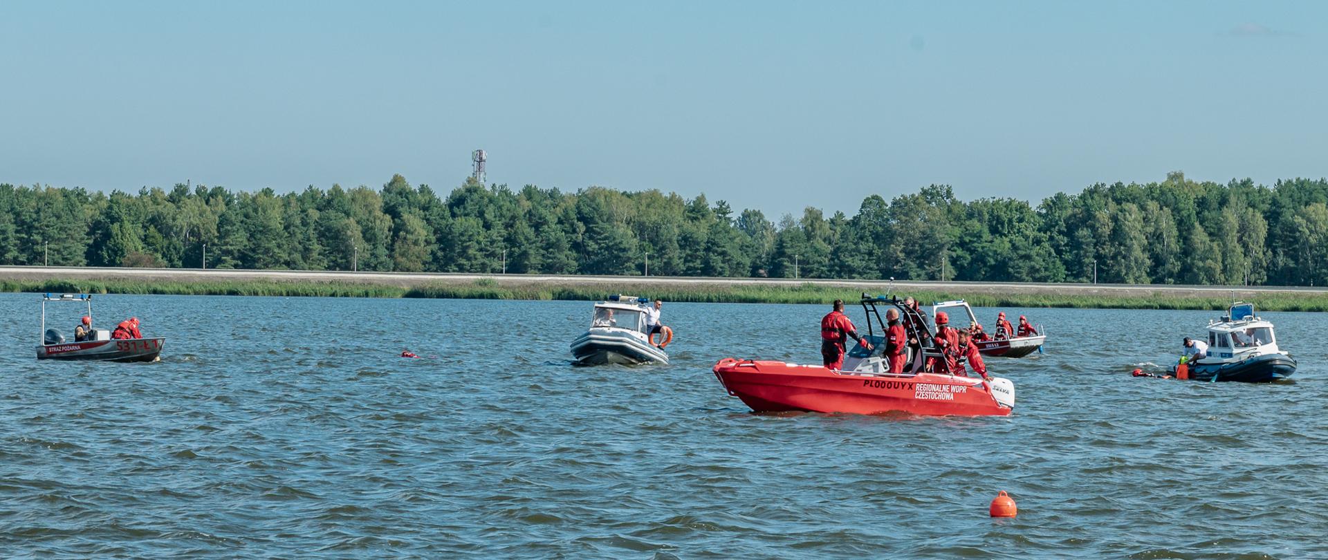 Zdjęcie przedstawia działania ratownicze prowadzone podczas ćwiczeń na Zalewie Porajskim. Strażacy oraz przedstawiciele służb współdziałają podczas ćwiczeń z zakresu ratownictwa wodnego oraz działań poszukiwawczo-ratowniczych na obszarach wodnych. Na zdjęciu widoczne są osoby uczestniczące w działaniach oraz sprzęt wykorzystywany do prowadzenia akcji ratowniczej na wodzie.