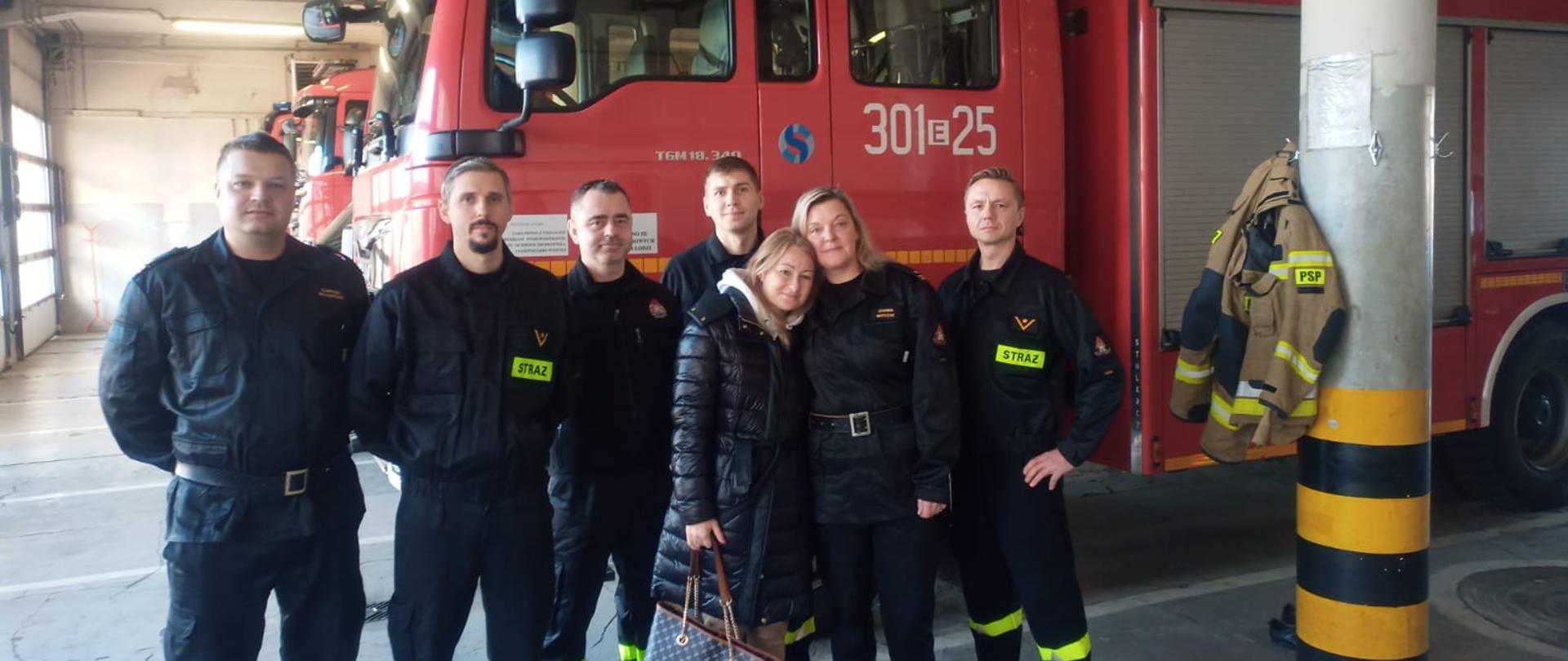 Strażacy stojący w rzędzie na tle samochodu pożarniczego. Wśród nich kobieta kobieta w czarnej kurtce z torbą w ręku