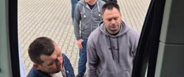 Widok z wnętrza pojazdu przez otwarte drzwi. Przed wejściem stoi sześciu mężczyzn ustawionych w kolejce na brukowanym podjeździe. W tle znajduje się jasny budynek z oknami oraz zadbana zieleń. Mężczyźni są ubrani głównie w bluzy i kurtki w stonowanych kolorach, a ich uwagę skupia wnętrze pojazdu.