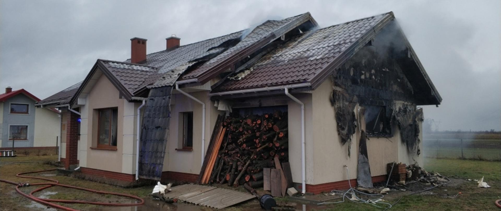 Na zdjęciu widać budynek, który się palił. Widać spaloną elewację ściany szczytowej oraz zerwaną blachę na dachu. Przed budynkiem rozwinięte linie wężowe.