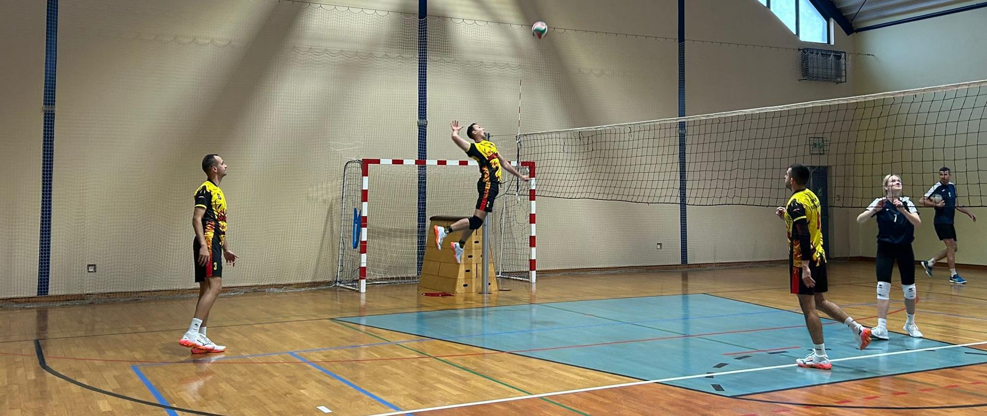 Dynamiczne ujęcie przedstawia zawodnika w wyskoku, który przygotowuje się do wykonania mocnego ataku nad siatką. Pozostali gracze na boisku czujnie obserwują lot piłki, zajmując pozycje defensywne.