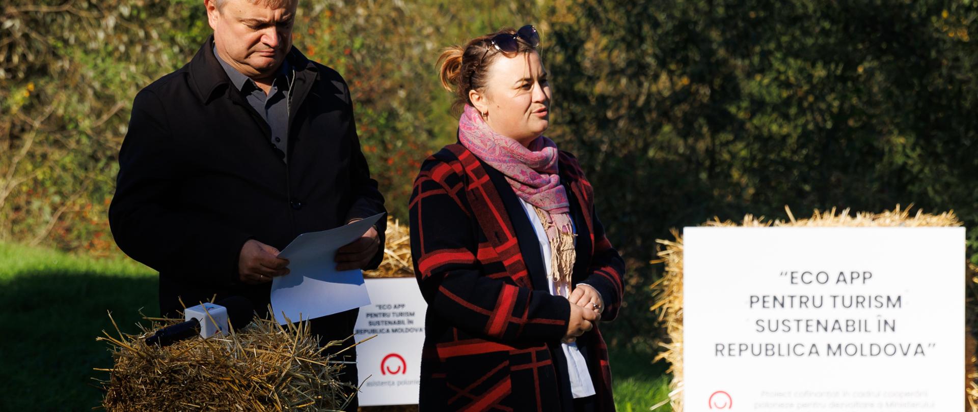 Zdjęcie przedstawia dwie osoby podczas uroczystości. Kobieta przemawia, mężczyzna sprawdza coś na kartce. W tle tablica informacyjna z logo polskiej pomocy 