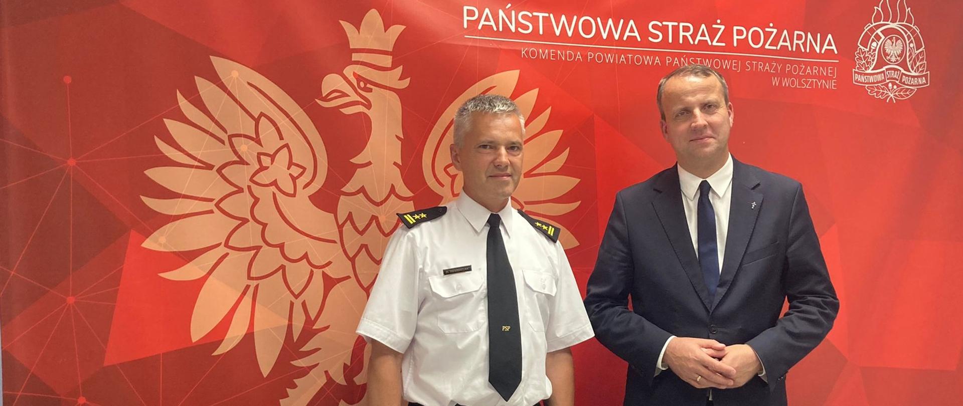 Na zdjęciu widoczny Wojewoda Wielkopolski Pan Michał Zieliński (po prawej) stoi wraz z Komendantem Powiatowym PSP w Wolsztynie bryg. Romanem Rembielakiem. W tle baner w kolorze czerwonym z symbolem orła oraz napisem Państwowa Straż Pożarna.