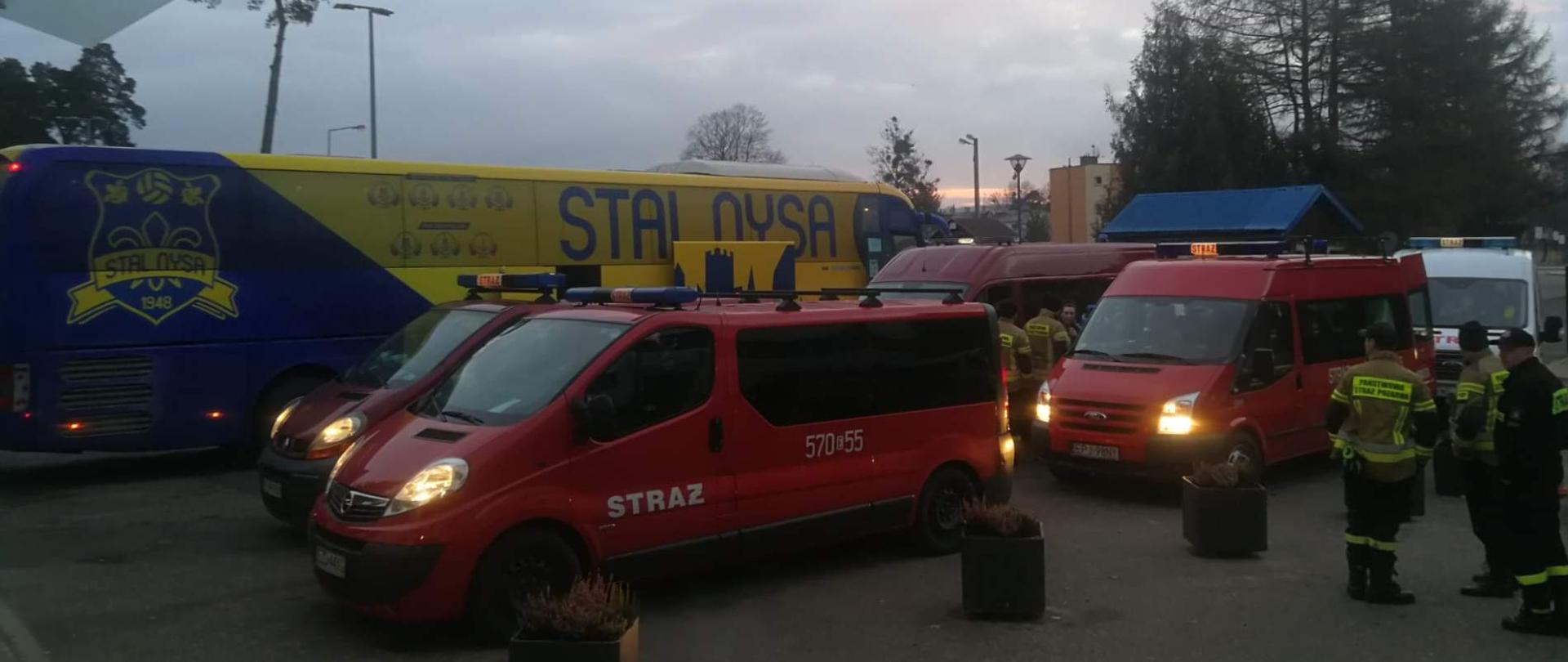 Na placu stoją samochody strażackie. Obok samochodów stoją strażacy. Za samochodami stoi duży żółto niebieski autokar. W tle widać szare niebo i drzewa.