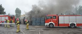Akcja na składowisku odpadów. Plac, na którym znajdują się pryzmy z odpadami. Na pierwszym planie dwa wozy straży pożarnej oraz strażacy prowadzący działania gaśnicze. Na ziemi znajdują się rozwinięte węże strażackie i rozdzielacz. Nad pryzmami unoszą się kłęby dymu. W tle drzewa. 