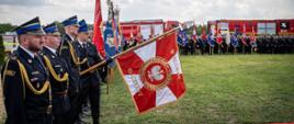 od lewej dwóch strażaków, następnie salutujący poczet sztandarowy komendy PSP, dalej stoją inne poczty sztandarowe a za nimi dwa samochody pożarnicze