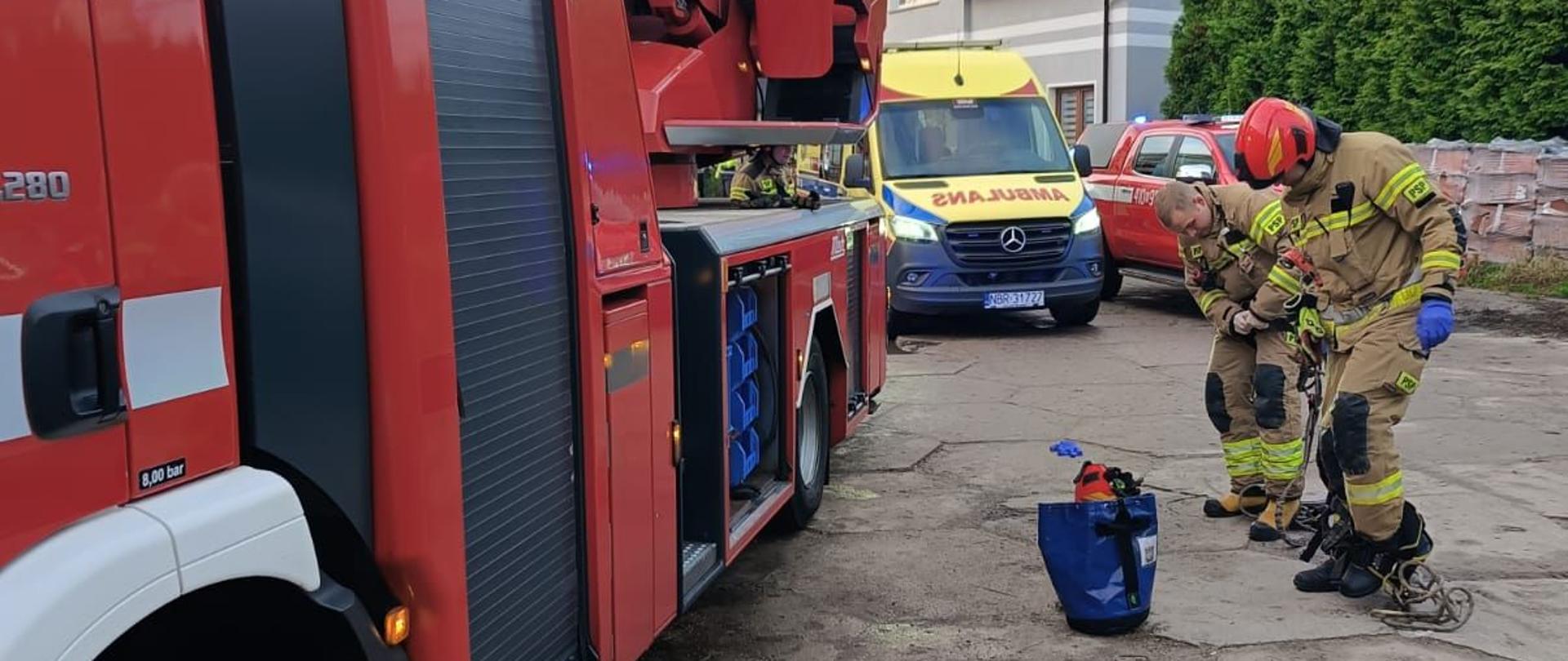 Na zdjęci z prawej strony dwóch strażaków ubiera się w specjalny sprzęt. Z lewej strony czerwony duży samochód strażacki.