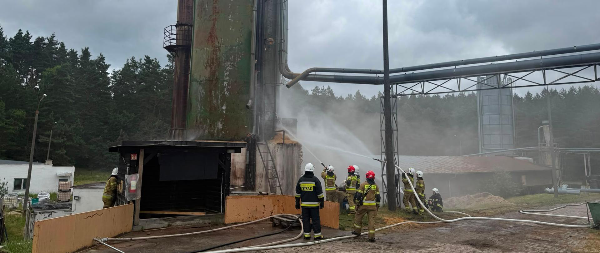 Grupa strażaków gasi pożar wysokiego, zielonego silosu trocin na terenie tartaku, kierując strumienie wody na konstrukcję pod zachmurzonym niebem