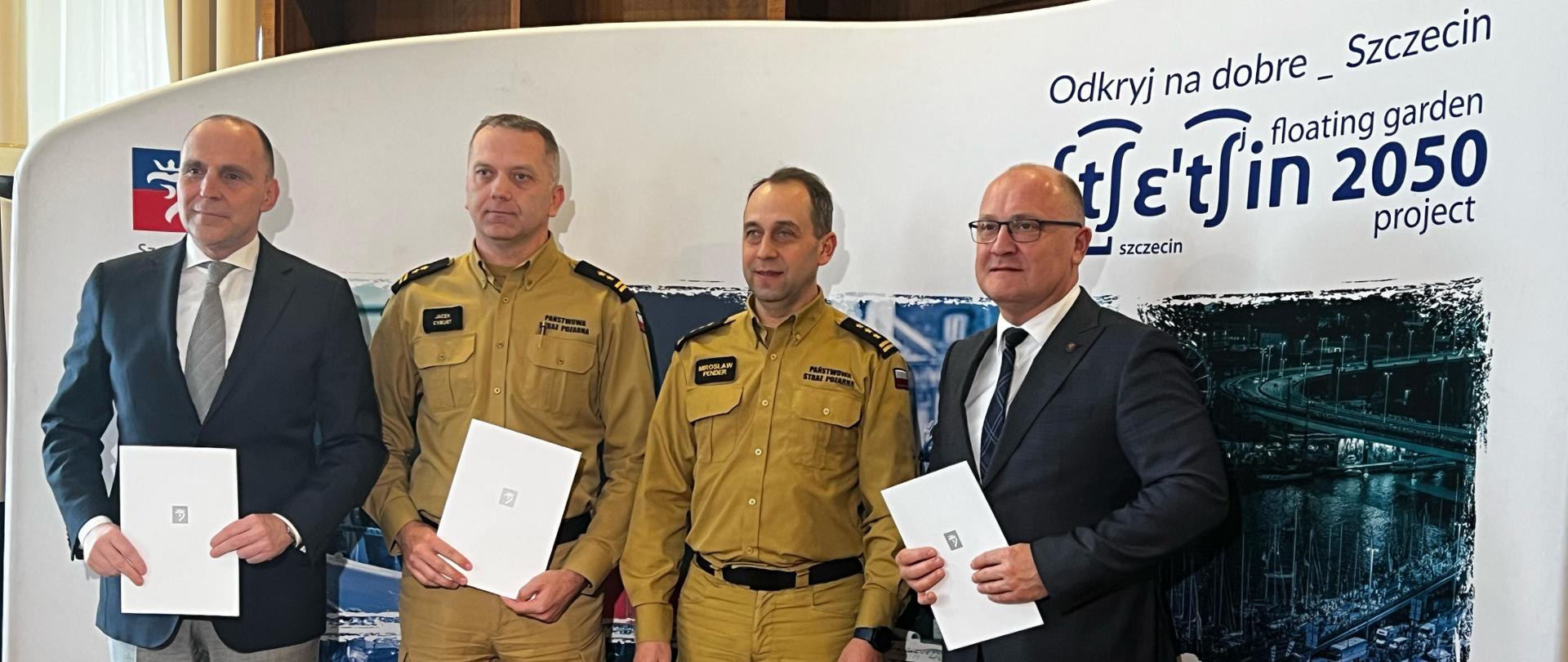 Na zdjęciu znajdują się cztery osoby stojące na tle banera promocyjnego miasta Szczecin z hasłem „Odkryj na dobre Szczecin Floating Garden 2050 Project”. Dwaj mężczyźni są ubrani w mundury strażackie, a dwaj pozostali w eleganckie garnitury. Każda z osób trzyma dokument, co sugeruje, że jest to moment podpisania lub ogłoszenia porozumienia czy umowy. W kadrze widać również mikrofon lokalnej telewizji TVP3 Szczecin. Pomieszczenie wygląda na oficjalne miejsce konferencji lub spotkania prasowego.