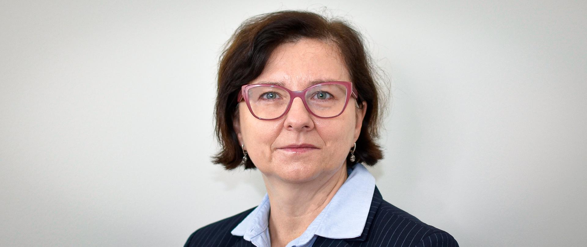 Kobieta w ciemnej marynarce w paski, jasnej bluzce i okularach, z krótkimi, ciemnymi włosami