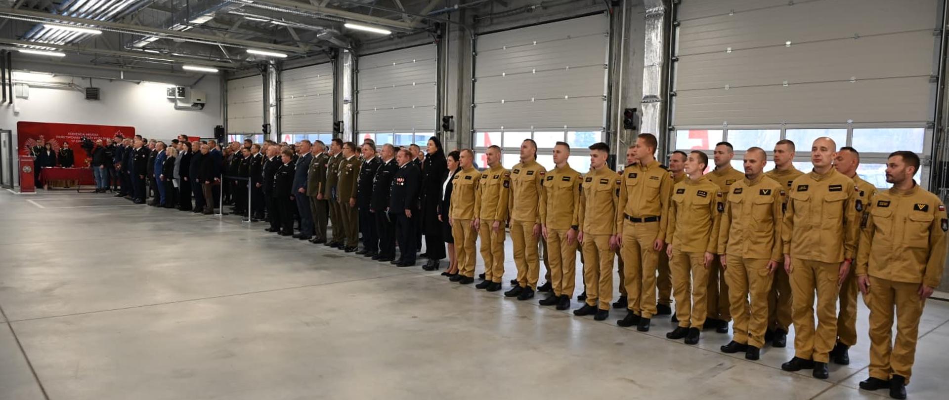 Strażacy stoją w szeregu na garażu jednostki.