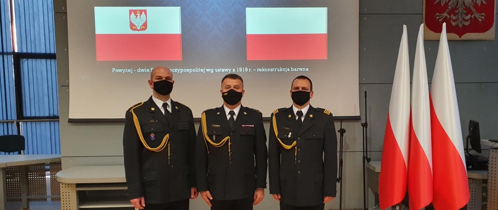 Na zdjęciu widoczni trzej strażacy w mundurach galowych koloru granatowego bez nakrycia głowy po prawej stronie za nimi trzy biało czerwone flagi za ich plecami napis uroczysta zbiórka z okazji Narodowego Święta Niepodległości