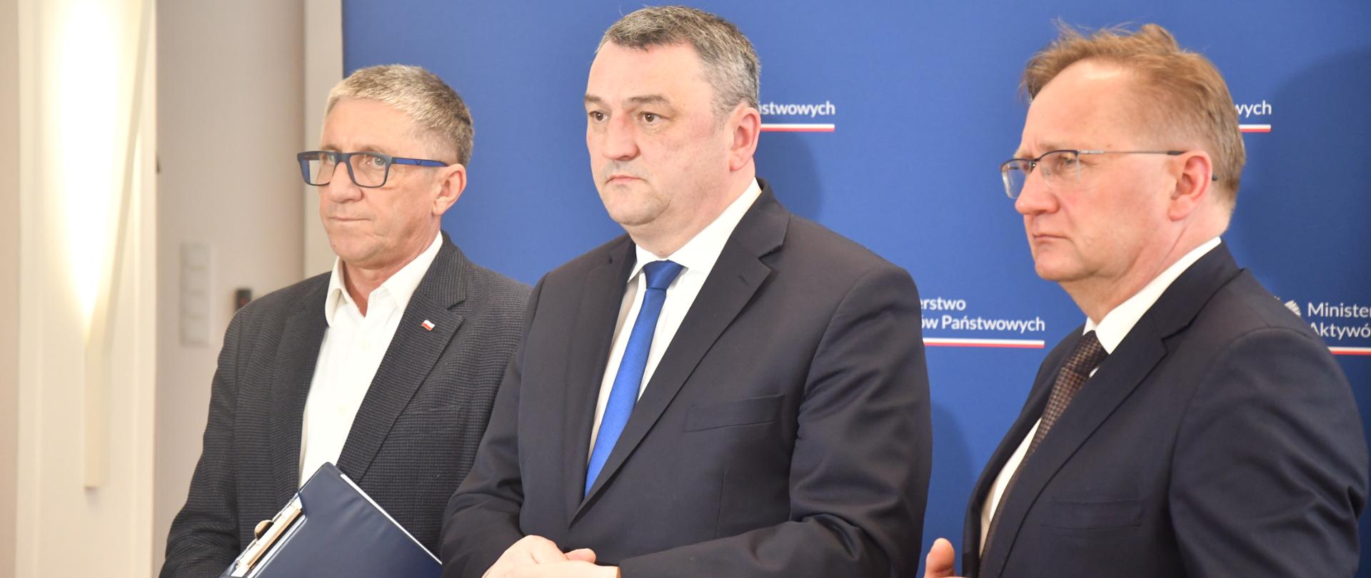 Wiceminister Marek Wesoły przemawia do mikrofonu. Obok stoją Poseł Grzegorz Matusiak oraz Poseł Andrzej Gawron. W tle ścianka z logotypami MAP oraz flaga unijna. Ujęcie zza kamery. 