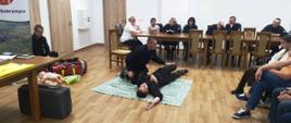 Strażacy prowadzą instruktarz udzielania pierwszej pomocy. Strażak pokazuje jak wygląda ułożenie osoby w pozycji bocznej bezpiecznej. W tle osoby przyglądające się prelekcji.