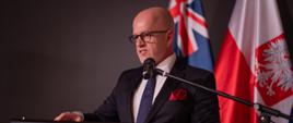 Konsul Generalny RP Piotr Rakowski wita zebranych gości przy mównicy na tle flag państwowych.