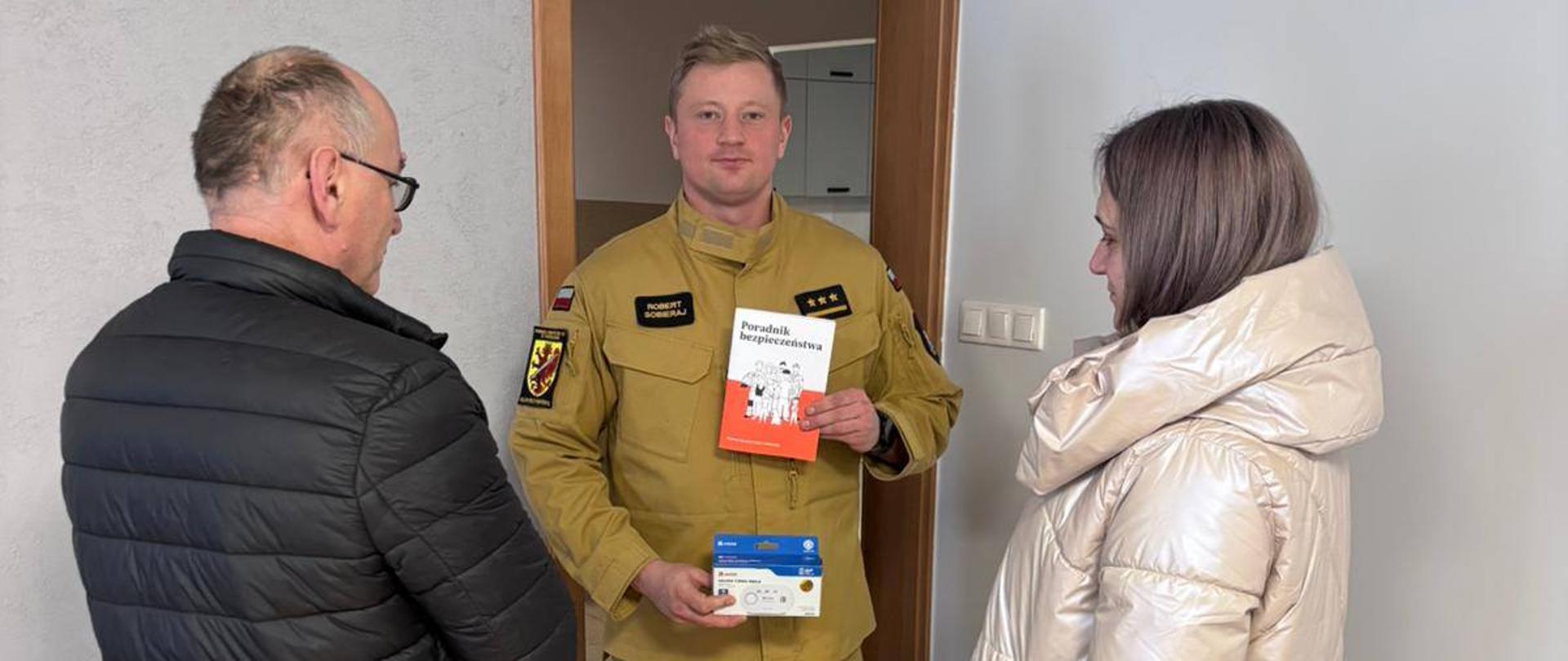 Strażak PSP trzyma czujkę dymu i tlenku węgla oraz poradnik, rozmawiając z dwojgiem dorosłych w pomieszczeniu biurowym.