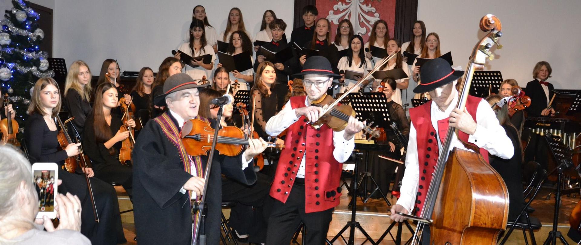 trzech góralskich muzyków grających na skrzypcach i kontrabasie na tle orkiestry symfonicznej i chóru na scenie w sali królewskiej psm mielec