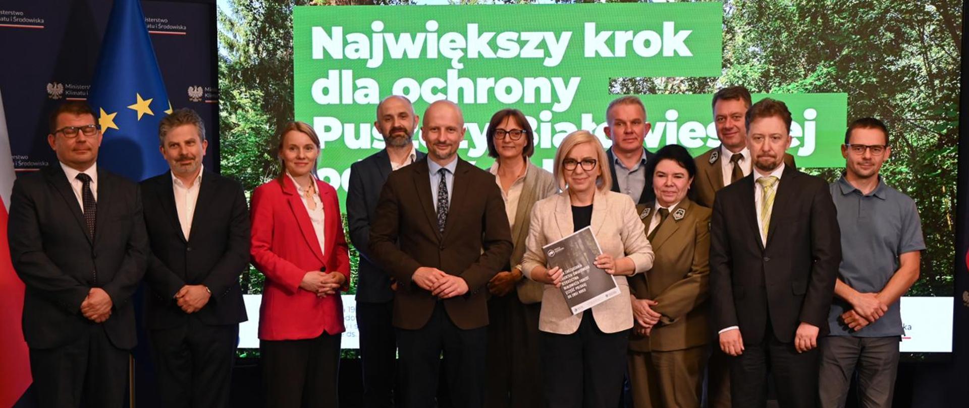 Minister of Climate and Environment Paulina Hennig-Kloska and Deputy Minister of Climate and Environment Mikołaj Dorożała during the ceremony marking the signing of the Management Plan for the Białowieża Forest World Heritage Site. The Minister and Deputy Minister pose for a group photograph with ten other participants. The Minister is holding the Management Plan for the Białowieża Forest World Heritage Site.