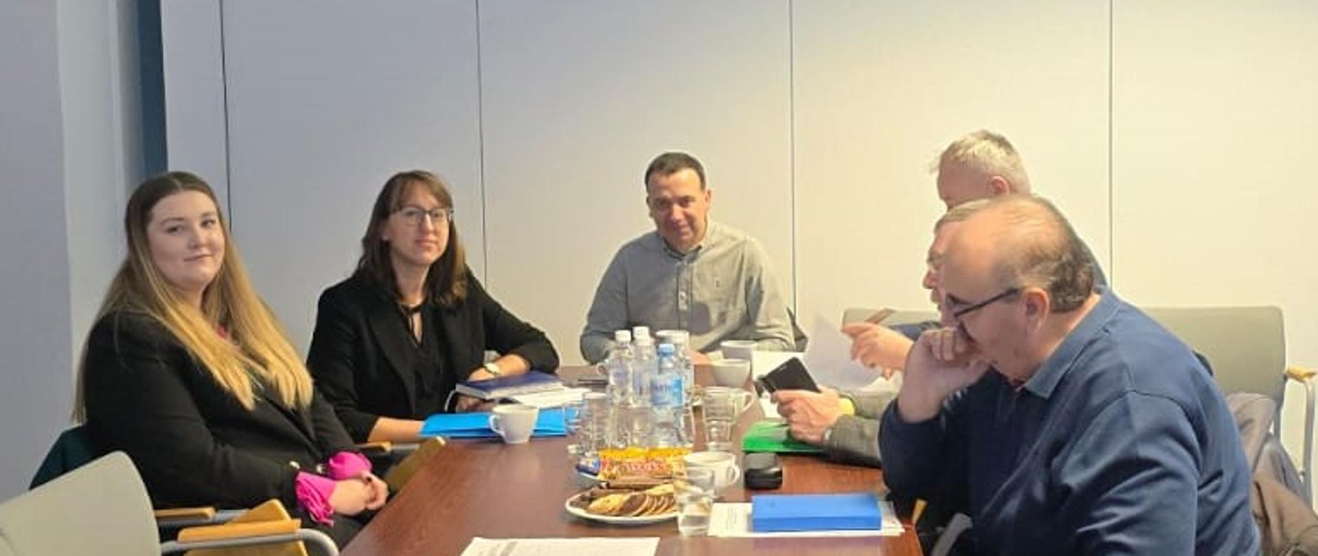 Na zdjęciu widać grupę pięciu osób (trzech mężczyzn i dwie kobiety) siedzących przy prostokątnym stole w jasnej sali konferencyjnej.
• Siedzący przy stole:
o Po lewej stronie, bliżej nas: Dwie kobiety w ciemnych ubraniach (jedna z nich się uśmiecha).
o Na wprost, w głębi: Mężczyzna w jasnoszarej koszuli.
o Po prawej stronie, bliżej nas: Dwóch mężczyzn, jeden z nich w granatowym swetrze lub bluzie, a drugi, częściowo zasłonięty, w jasnej koszuli. Mężczyzna w granatowym swetrze opiera brodę na dłoni, wydaje się słuchać lub zastanawiać.
• Na stole widać butelki z wodą, szklanki, dokumenty w niebieskich teczkach, oraz talerz z przekąskami (np. pączkami lub ciastkami).
• Pomieszczenie:
o Ściany są białe. Na jednej ze ścian wiszą głośniki i znajduje się jasne, drewniane drzwi.
o Na suficie zamontowane są lampy rastrowe oraz projektor, który wyświetla niebieskie światło.
o Po prawej stronie widoczne są wysokie, jasnoniebieskie/szare żaluzje pionowe.
o Podłoga pokryta jest ciemną, niebieską wykładziną.
o Wokół stołu oraz na pierwszym planie znajdują się puste krzesła konferencyjne z odchylanymi pulpitami.
Zdjęcie przedstawia spotkanie lub naradę w sali konferencyjnej.
