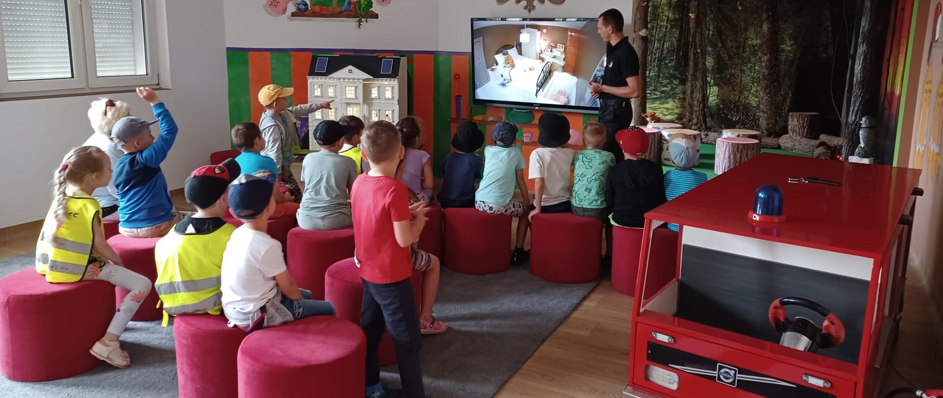 Na zdjęciu grupa dzieci słucha prezentacji na Sali edukacyjnej „Ognik”, którą prowadzi strażak PSP. Na telewizorze wyświetla się obraz z kamery umieszczonej w makiecie domu jednorodzinnego, po prawej stronie wóz strażacki dziecięcych rozmiarów.