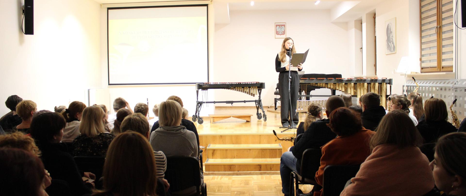 publiczność na sali koncertowej słucha osoby zapowiadającej kolejnego wykonawcy