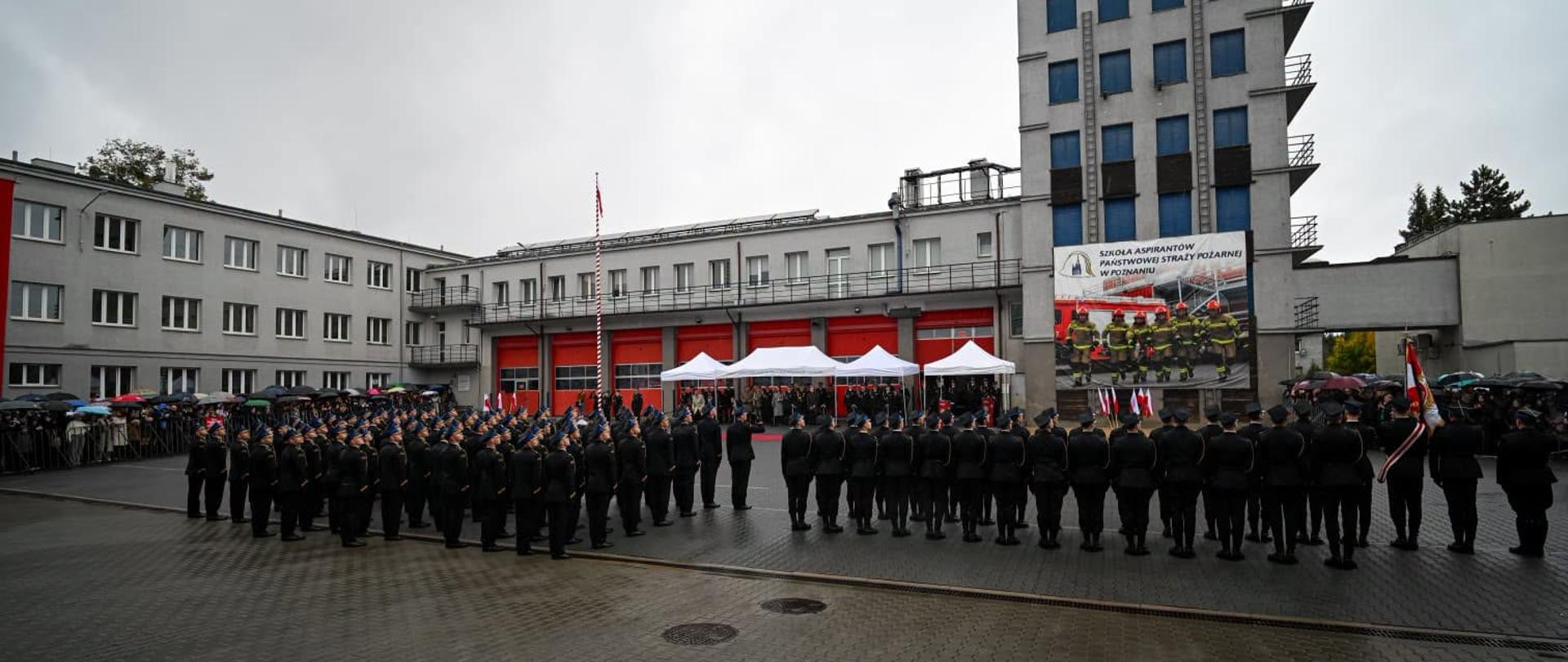 Strażacy, którzy złożą za chwilę ślubowanie, są ustawieni w pododdziałach na placu/dziedzińcu szkoły aspirantów. Wokół zebrani goście, pada deszcz.