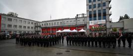 Strażacy, którzy złożą za chwilę ślubowanie, są ustawieni w pododdziałach na placu/dziedzińcu szkoły aspirantów. Wokół zebrani goście, pada deszcz.