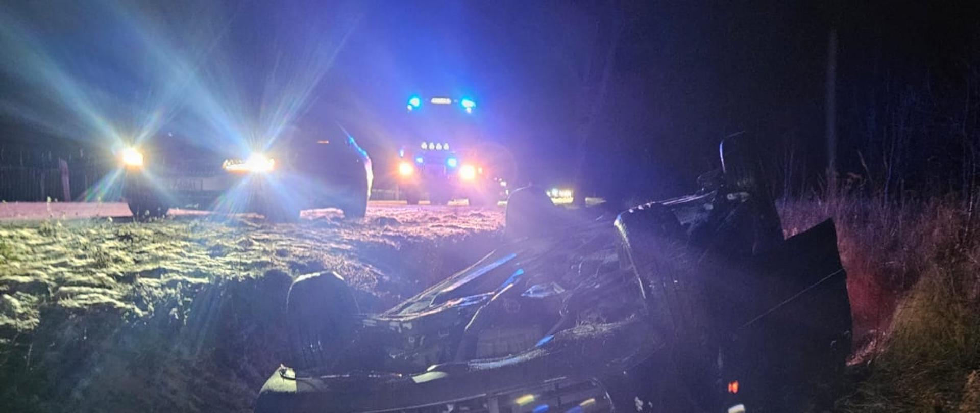 Noc. Samochód osobowy leży na dachu w rowie. Z lewej na drodze stoi samochód strażacki, niebieskie światła.