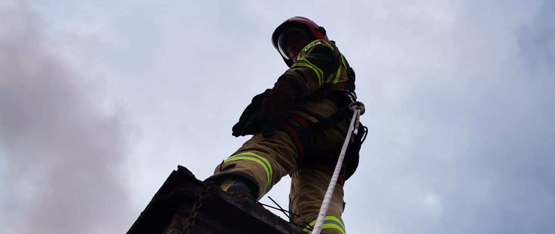 Strażak gasi pożar sadzy w kominie - stoi ba kominie zapięty linką