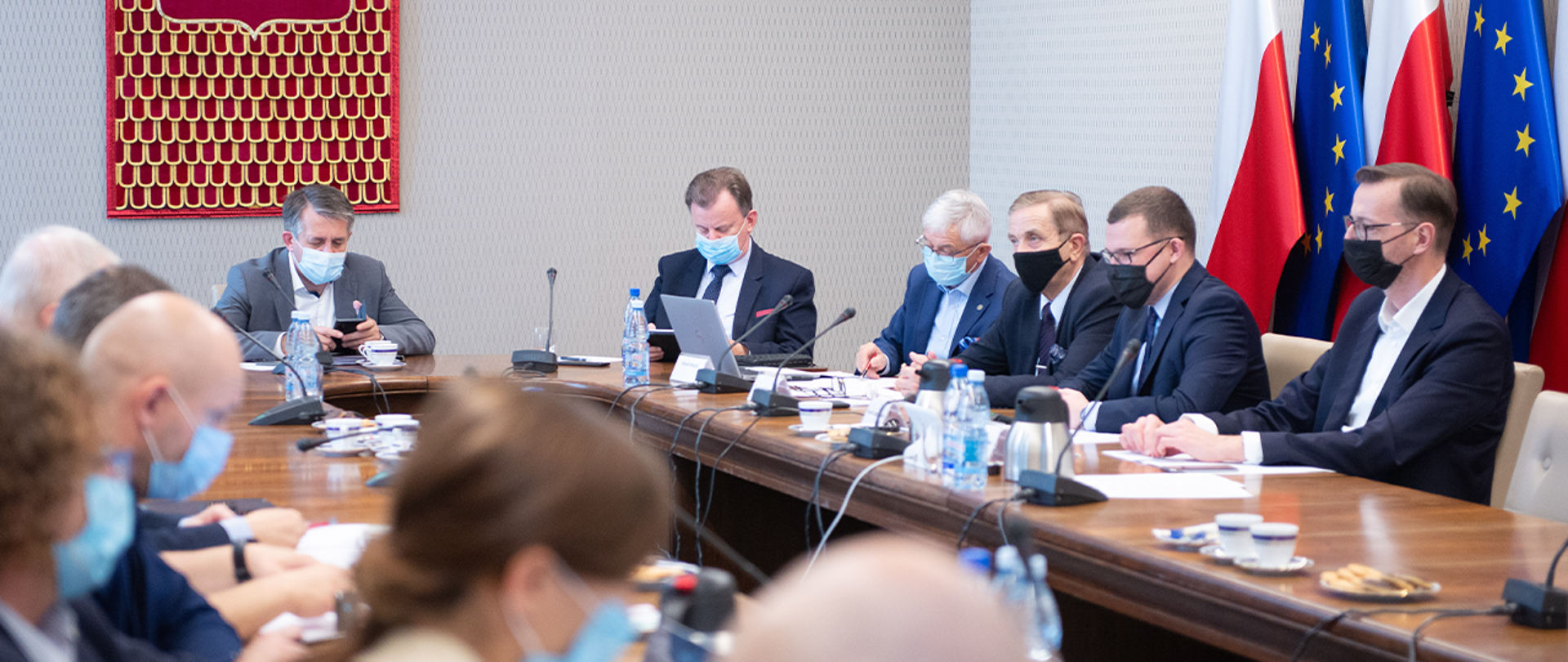 Zdjęcie przedstawia wiceministra Pawła Szefernakera wraz z innymi uczestnikami w trakcie obrad Komisji Wspólnej Rządu i Samorządu Terytorialnego. W tle znajdują się flagi Polski i Unii Europejskiej.