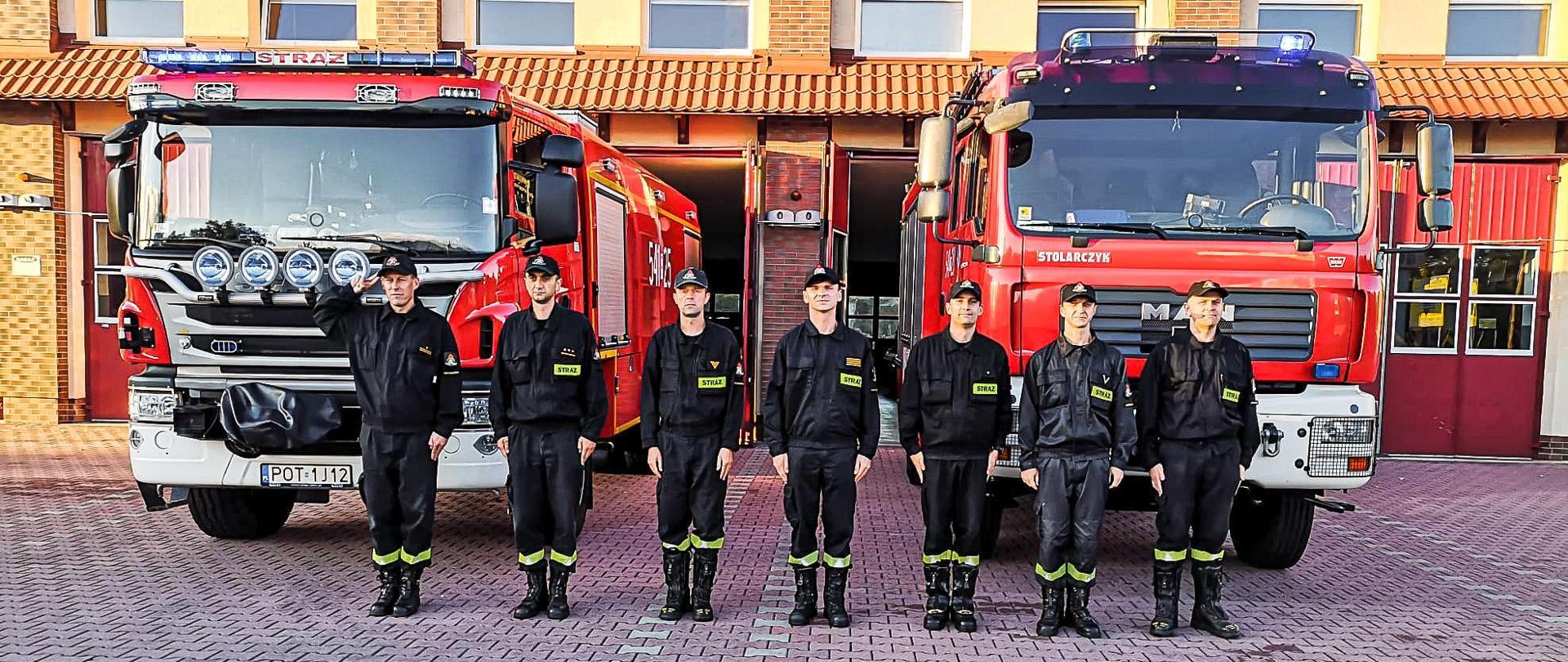 Strażacy w ubraniach koszarowych stoją przed samochodami strażackimi. W tle budynek straży pożarnej.